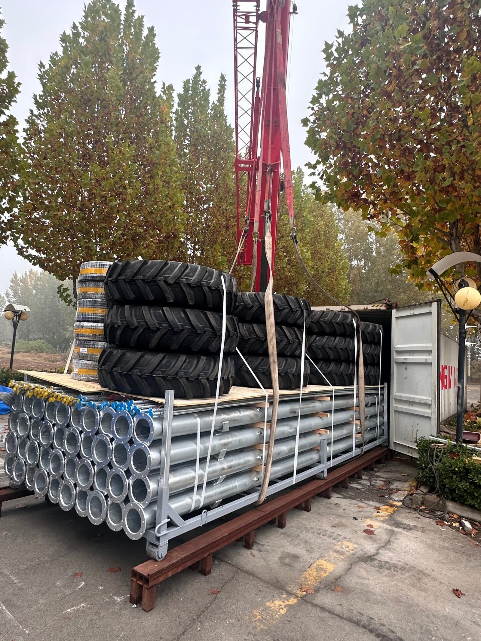 A red crane lifts a flatbed lorry loaded with stacks of large tractor tyres and metal pipes.