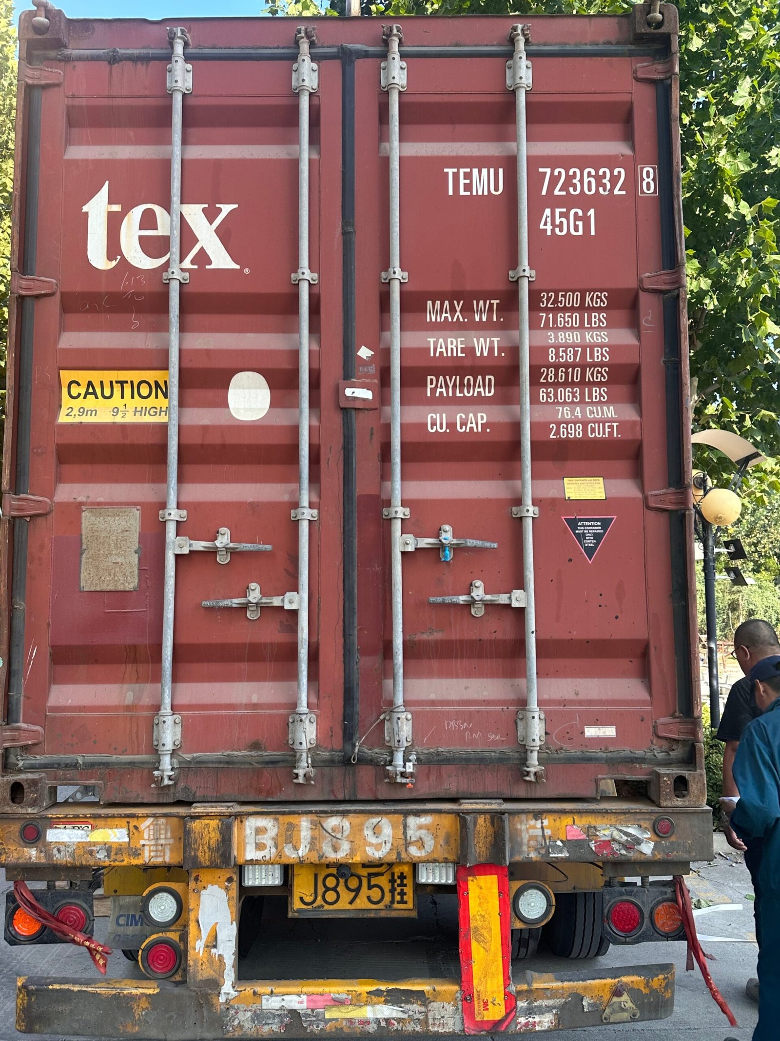 The back of a red Tex shipping container with weight specifications printed on the door, mounted on a weathered yellow lorry bed.