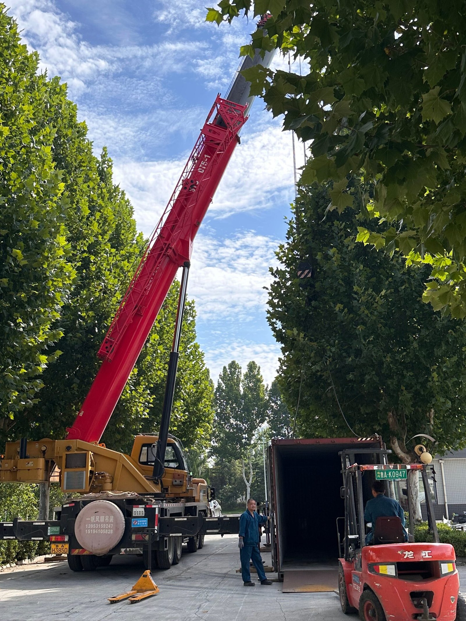 A red mobile crane lifting a shipping container, with a forklift and two workers nearby.