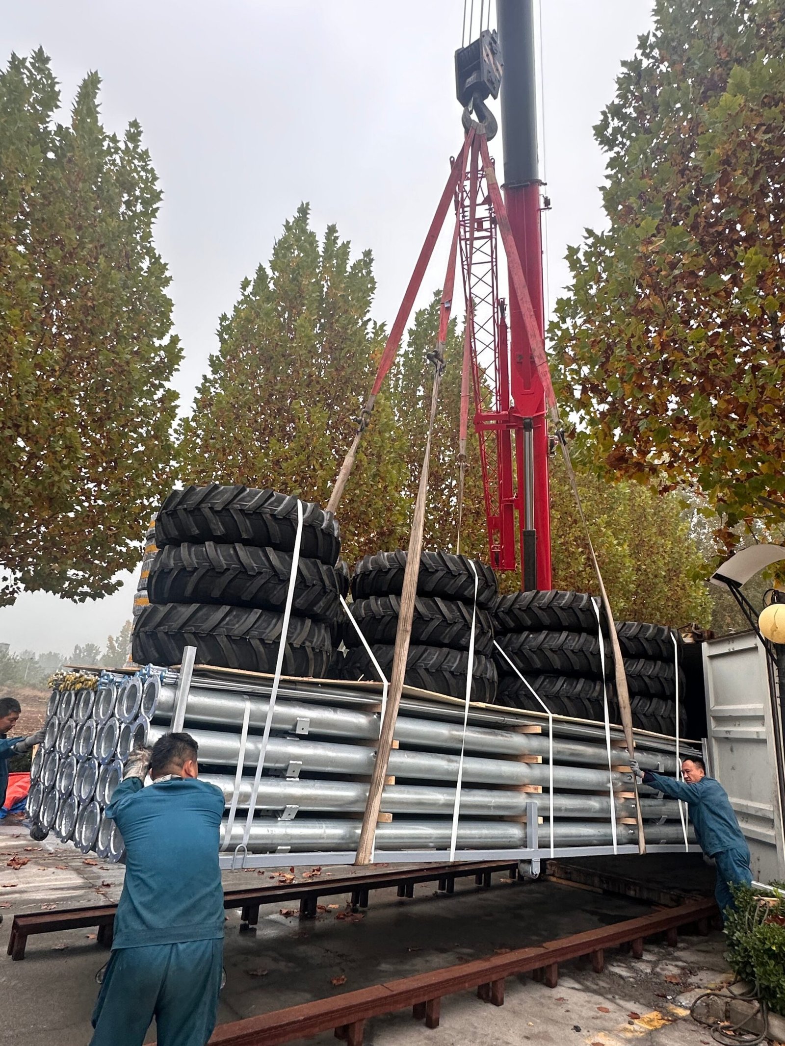 Two workers guide a crane lifting a metal pallet loaded with steel pipes and large tyres onto a lorry bed.